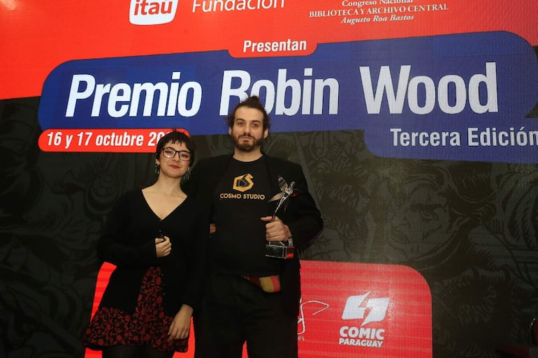 Mujer joven con blusa negra y gafas, junto a hombre en camiseta negra con trofeo, en evento de premiación con cartelero de fondo.