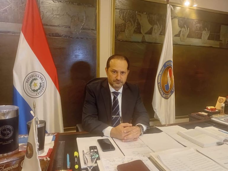Jalil Rachid, vestido con traje oscuro y corbata a rayas, serio, sentado en su oficina con documentos y teléfono móvil sobre el escritorio.
