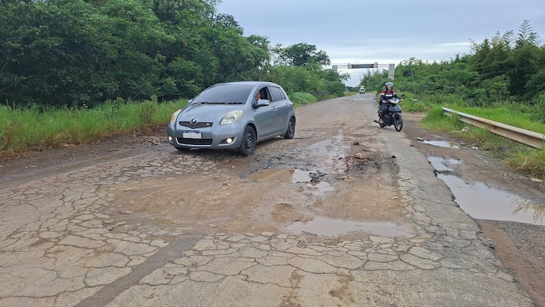 Vehículo plateado detenido junto a motociclista con casco, rodeados de baches y barro en una carretera deteriorada.