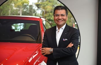 Hombre sonriente en traje oscuro y gafas, junto a un coche rojo, con un pin de fútbol en la solapa.
