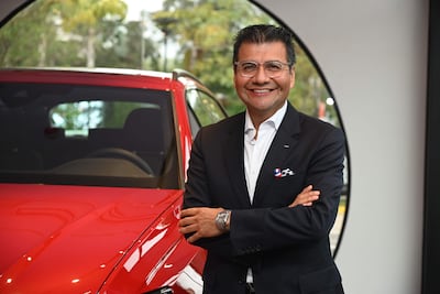 Hombre sonriente en traje oscuro y gafas, junto a un coche rojo, con un pin de fútbol en la solapa.