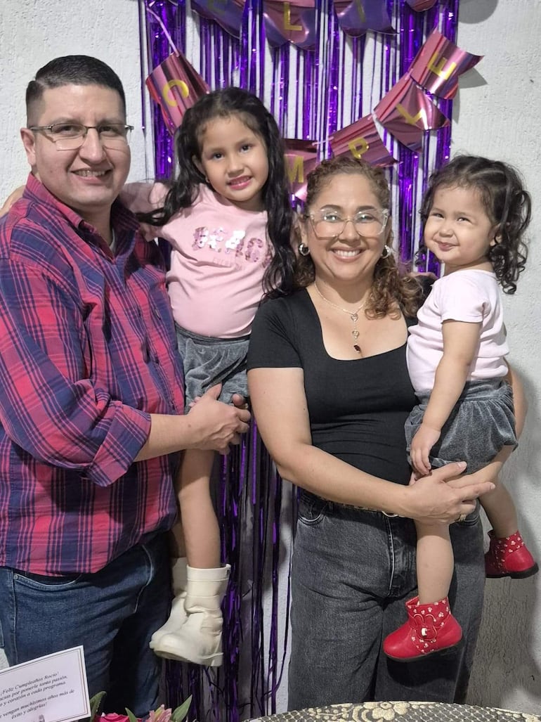 Mujer en camiseta negra abraza a niña con blusa rosa, mientras hombre con camisa de cuadros sostiene a otra niña, con decoraciones moradas al fondo.