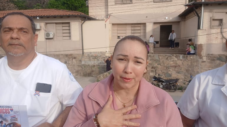 Mujer gesticulando con seriedad, acompañada de dos hombres con camisas blancas, en ambiente exterior de un edificio institucional.