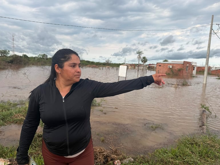 Mujer con chaqueta negra y leggings marrones, de pie en un área inundada, señalando hacia el fondo en un clima nublado.