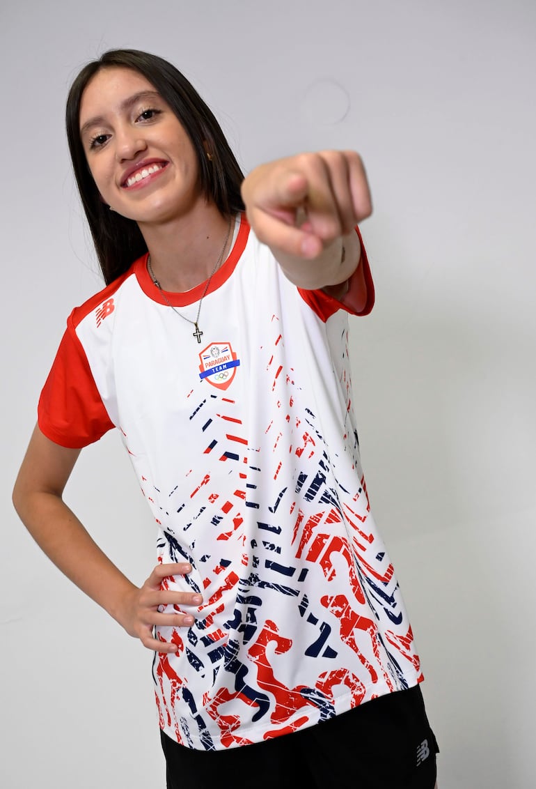 Mujer sonriente en camiseta blanca con detalles rojos y azules, apuntando a la cámara y sosteniéndose la cadera.