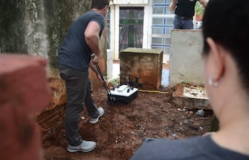 Hombre de espaldas con palanca, otro tomando notas y mujer observando, en un área de excavación al aire libre.