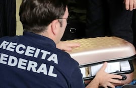 Funcionario de Receita Federal con camiseta oscura revisando un maletín abierto en ambiente cerrado.