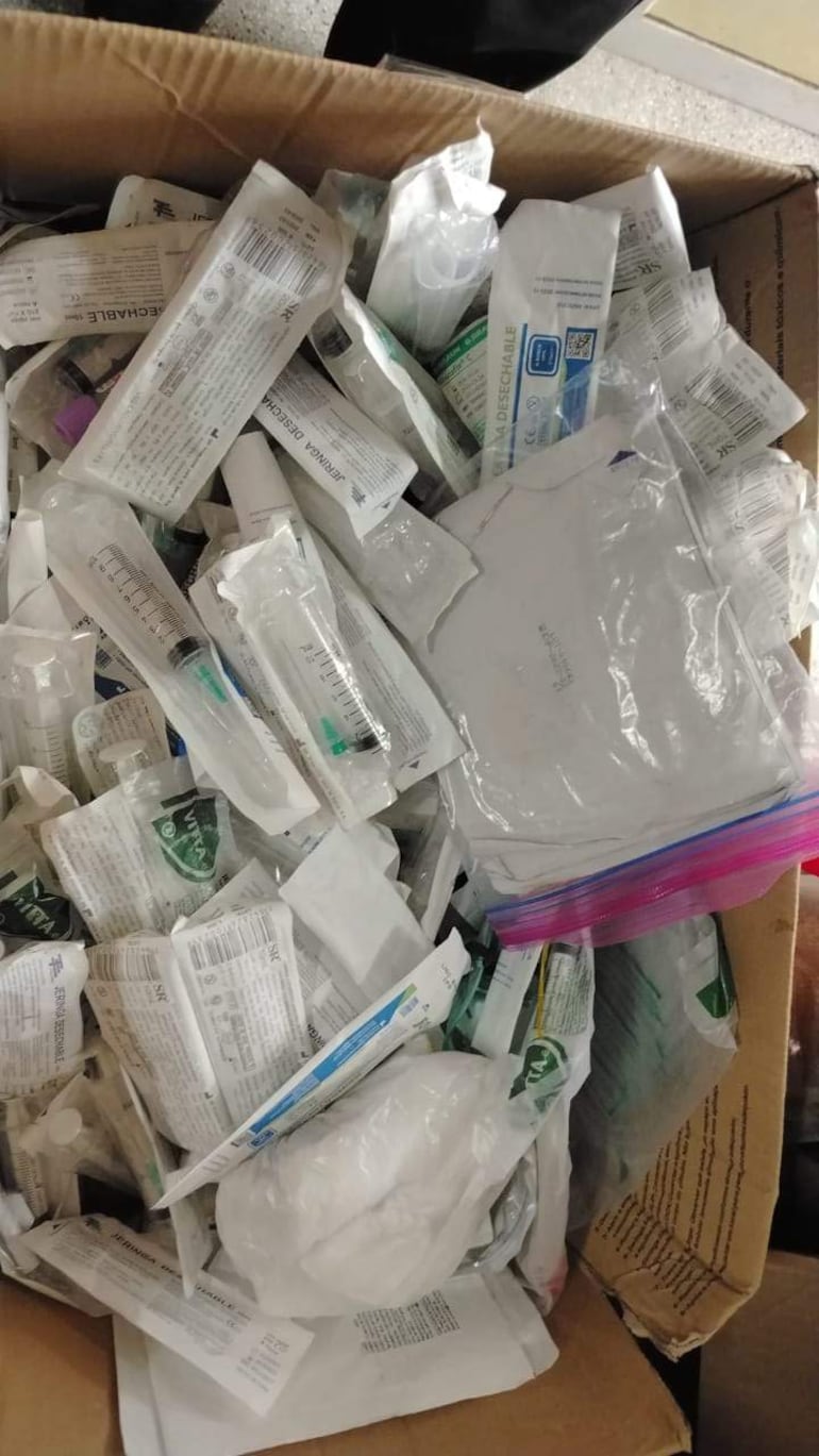 Caja de cartón con insumos médicos como jeringas y guantes desechables, apilados desorganizados en un ambiente interior.