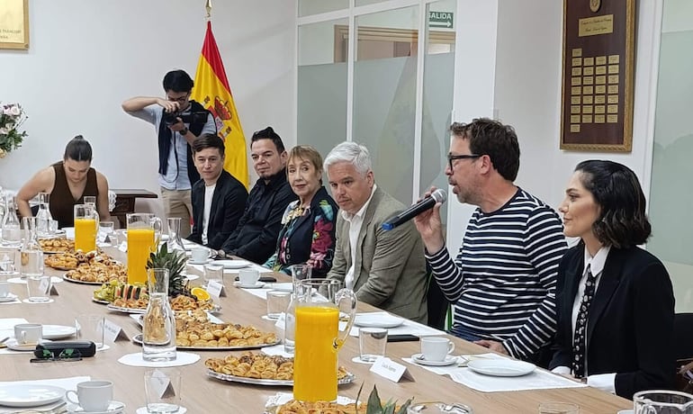 Martinessi en camiseta a rayas habla mientras Irún y Mora lo escuchan, rodeados de un ambiente formal y comensales atentos.
