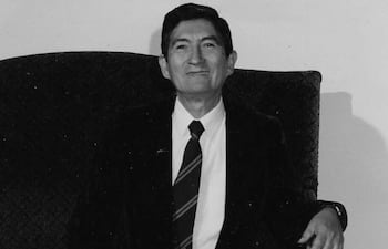 Narciso R. Colmán (h) sonriente en sillón, vestido con traje oscuro y corbata a rayas en un fondo neutro.