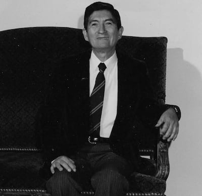 Narciso R. Colmán (h) sonriente en sillón, vestido con traje oscuro y corbata a rayas en un fondo neutro.