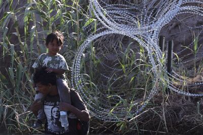 Un migrante con un niño en hombros camina a lo largo del Río Grande, bordeado con alambres de púa, en Eagle Pass, Texas.
