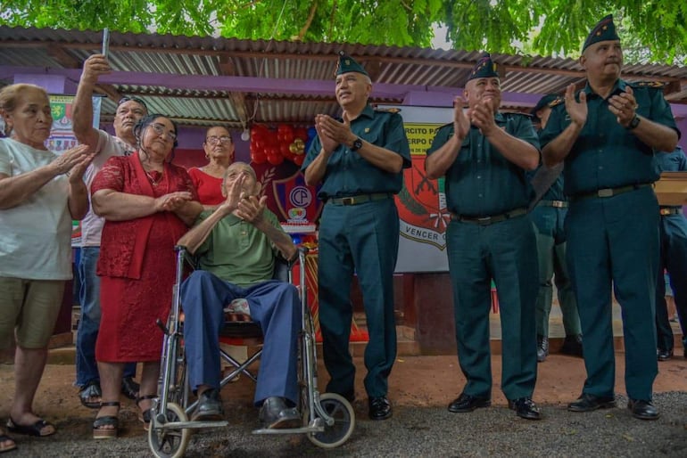 Cuatro personas aplauden, incluyendo a un anciano en silla de ruedas, mientras policías observan atentamente en un ambiente decorado.