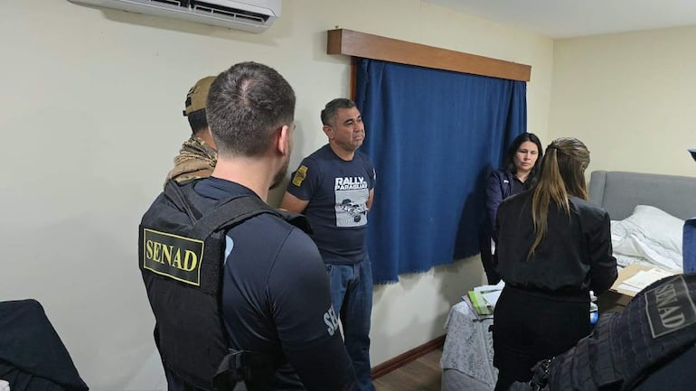 Hombre con camiseta azul oscuro del Rally Paraguay es interrogado por agentes de SENAD, en una habitación con documentos en la mesa.