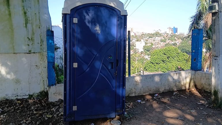 Baño portátil azul en área exterior con vegetación al fondo, sin personas visibles. Estructuras urbanas presentes.
