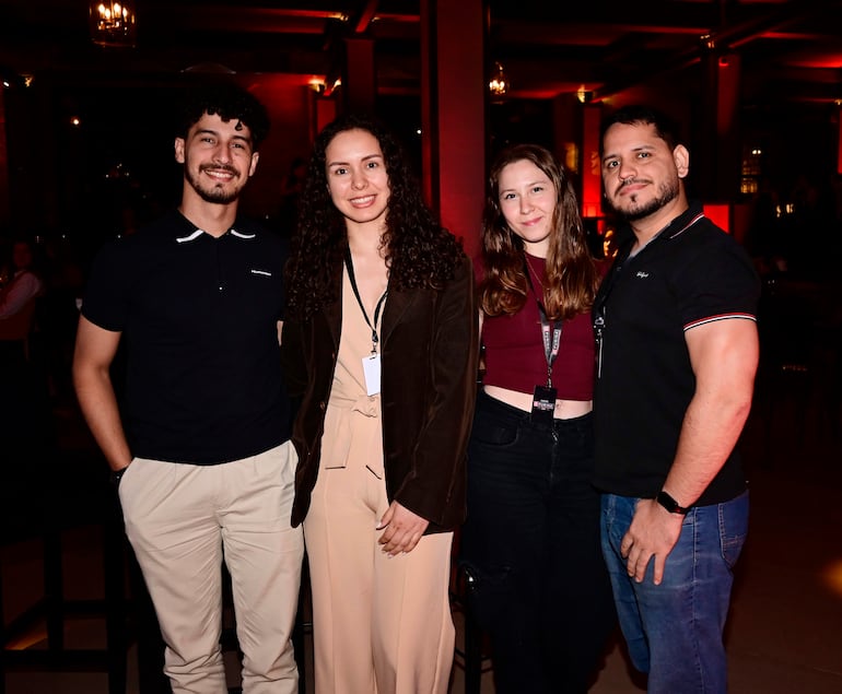 Hombre con cabello rizado y barba, mujer con atuendo marrón, mujer en top rojo, hombre con camiseta negra, todos sonriendo en evento festivo.