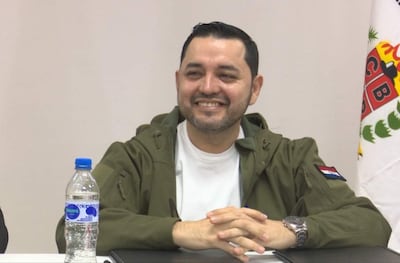 Luis Bello, con camiseta blanca y chaqueta verde, sonríe con las manos entrelazadas frente a una botella.