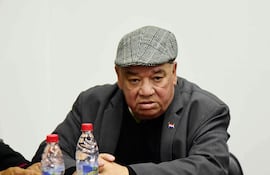 Hombre serio con abrigo oscuro y gorra, sentado en una mesa con botellas de agua, en ambiente neutral.