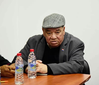 Hombre serio con abrigo oscuro y gorra, sentado en una mesa con botellas de agua, en ambiente neutral.