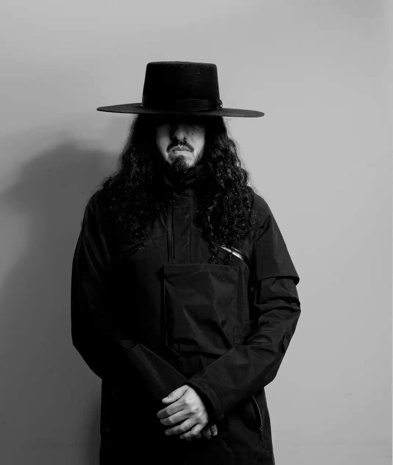 Hombre con cabello rizado y largo, vistiendo abrigo negro y sombrero, de pie con una postura tranquila.