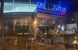 Fachada del Cosmopolitan Club iluminada, sin personas visibles y una motocicleta amarilla en la esquina inferior derecha.