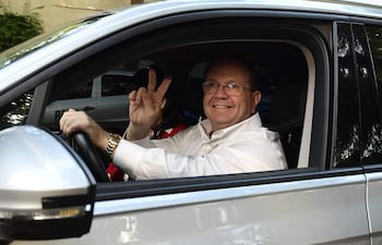 Hombre en camisa blanca y reloj dorado sonríe y hace un gesto de paz desde el coche. Mujer con blusa roja visible a su lado.