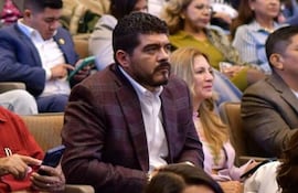 Hombre de cabello oscuro y barba, vestido con camisa blanca y saco marrón, escucha atentamente en evento rodeado de asistentes.