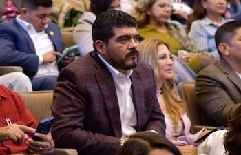 Hombre de cabello oscuro y barba, vestido con camisa blanca y saco marrón, escucha atentamente en evento rodeado de asistentes.