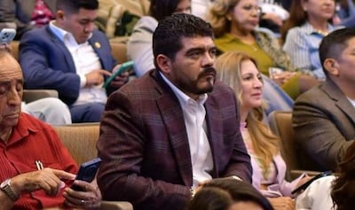 Hombre de cabello oscuro y barba, vestido con camisa blanca y saco marrón, escucha atentamente en evento rodeado de asistentes.