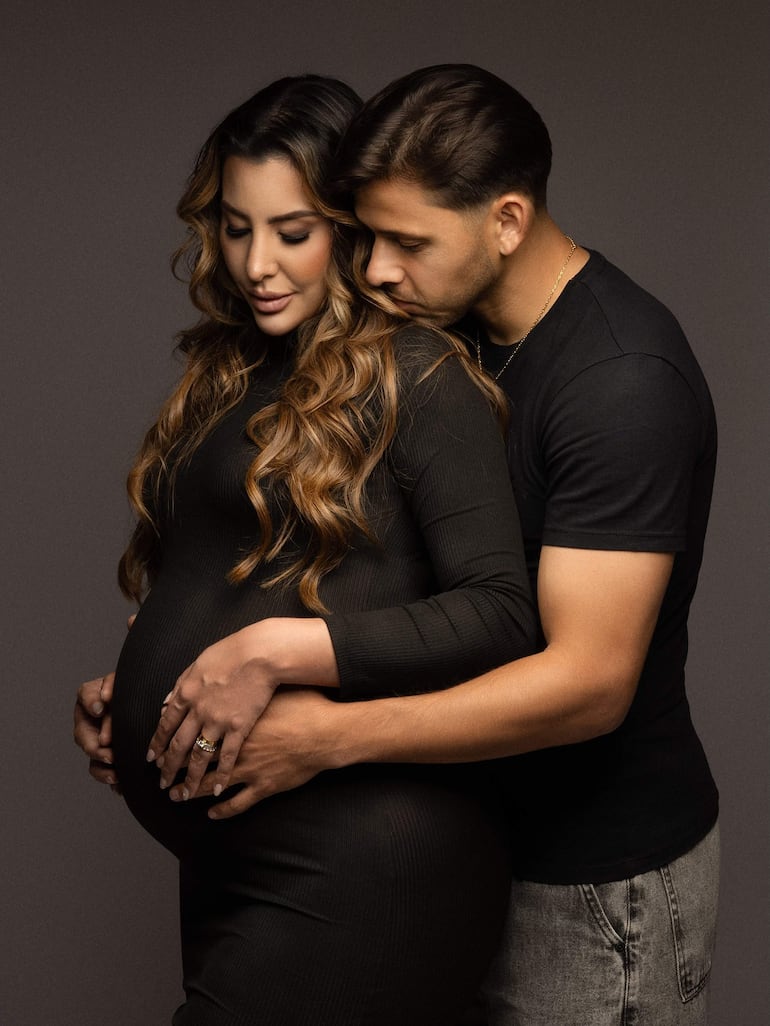 Larissa Riquelme, embarazada, en vestido negro ajustado, junto a Ángel Romero en camiseta negra, abrazándose afectuosamente.