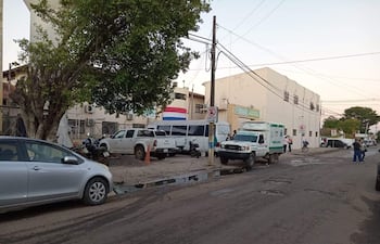 Calle urbana con baches y agua servida, ambulancia estacionada y un árbol frondoso a la izquierda.