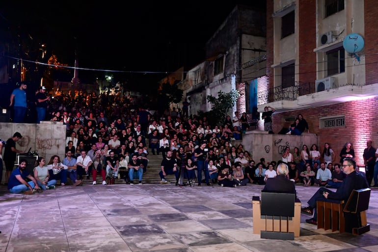 Hombres sentados, uno de espaldas y el otro hablando. Multitud atenta en un ambiente oscuro y iluminado.