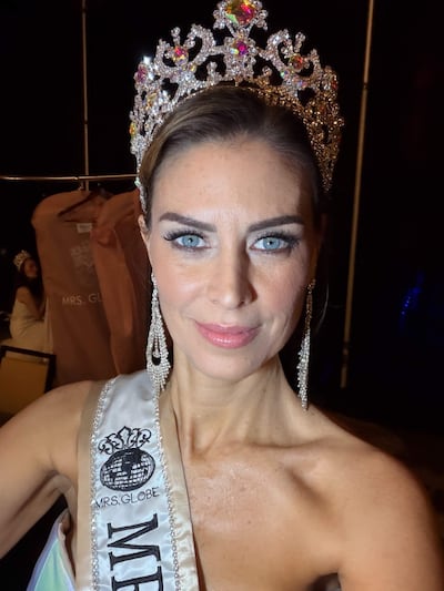 Mujer con corona brillante y banda MRS. GLOBE sonríe en backstage, con vestimentas colgadas en el fondo.