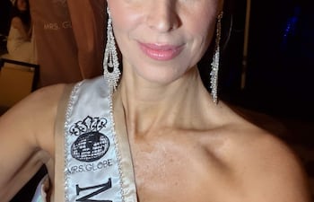 Mujer con corona brillante y banda MRS. GLOBE sonríe en backstage, con vestimentas colgadas en el fondo.