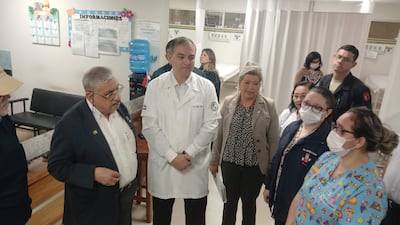 Un hombre con bata blanca, posiblemente médico, habla en el hospital rodeado de personas, algunas con mascarillas, en un ambiente iluminado.