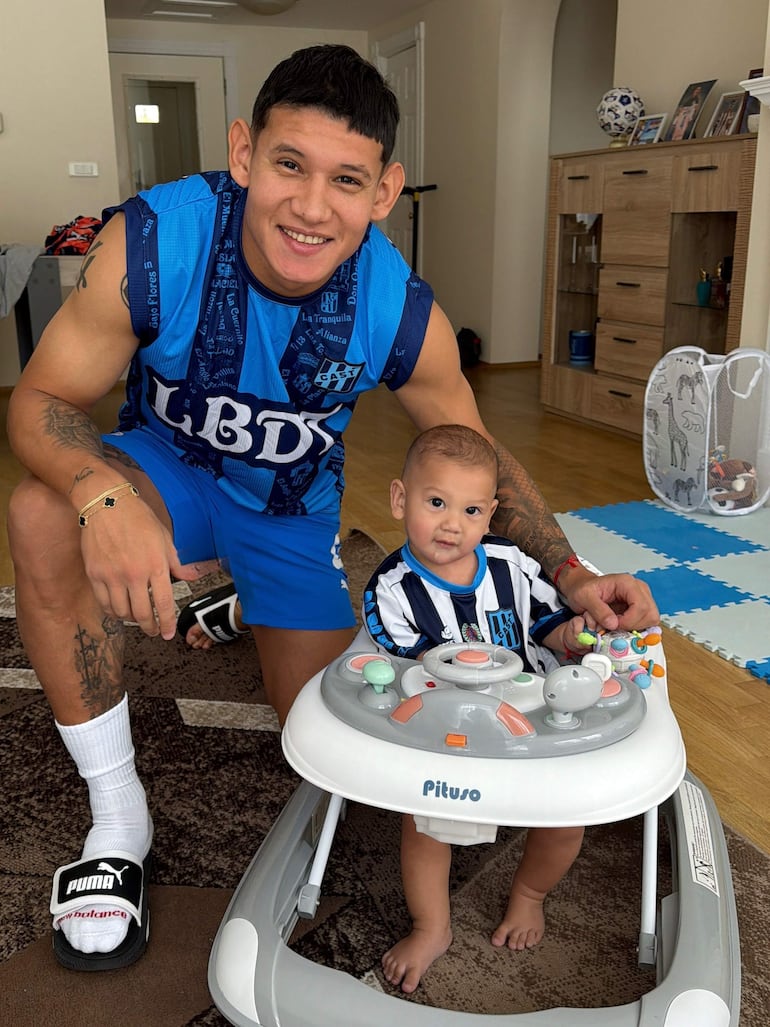 Juan Cáceres, con camiseta sin mangas azul, sonríe mientras se agacha junto a un niño en andador, en un ambiente decorado.