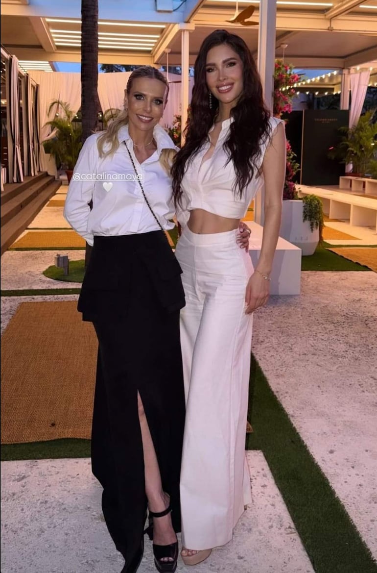 Gretha Matiauda y otra mujer sonríen, ambas con blusas blancas; se encuentran en un entorno decorado con plantas y luces.