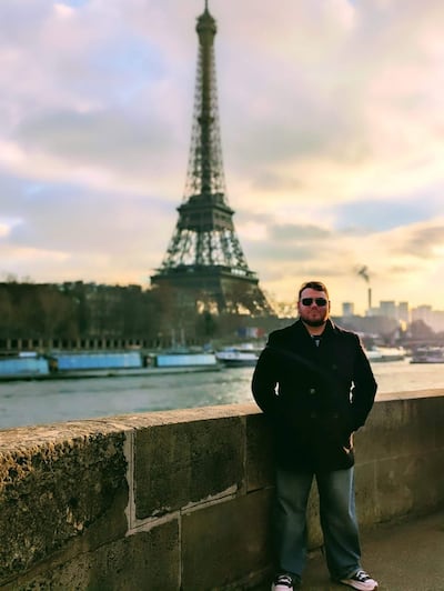 Hombre con abrigo oscuro y gafas de sol, de pie frente a la Torre Eiffel con los brazos en los bolsillos y el río Sena al fondo.