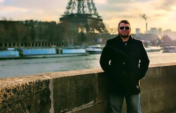 Hombre con abrigo oscuro y gafas de sol, de pie frente a la Torre Eiffel con los brazos en los bolsillos y el río Sena al fondo.