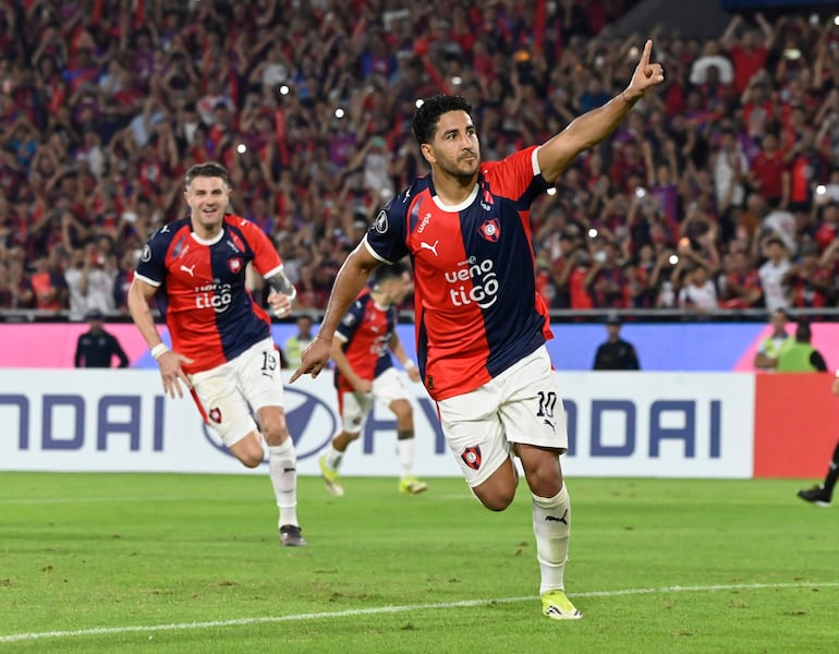 Jugador de Cerro Porteño con camiseta roja y azul, levantando un dedo en celebración, acompañado por un compañero en un estadio lleno.