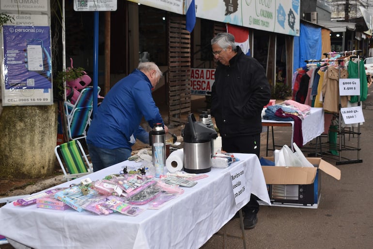 Hombre en chaqueta azul agachado sobre mesa con juguetes, mientras otro observa con abrigo negro en ambiente de feria.