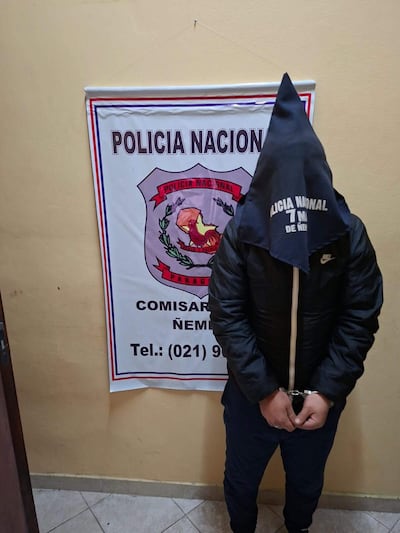Hombre con capucha y manos esposadas, delante de un cartel de la Policía Nacional en un ambiente de comisaría.