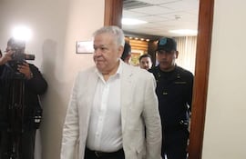 Jorge Brítez, vestido con traje claro y una camisa blanca, camina hacia una oficina junto a un agente de seguridad uniformado.