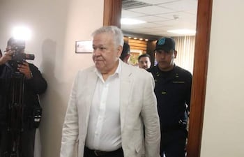 Jorge Brítez, vestido con traje claro y una camisa blanca, camina hacia una oficina junto a un agente de seguridad uniformado.