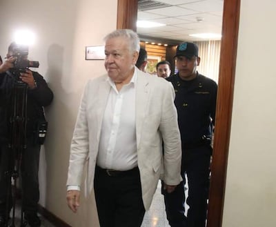 Jorge Brítez, vestido con traje claro y una camisa blanca, camina hacia una oficina junto a un agente de seguridad uniformado.