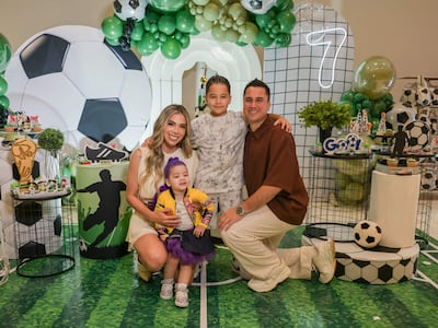 Mujer con vestido claro sostiene a una niña, mientras hombre y niño sonríen agachados en un entorno decorado con globos verdes y negros.