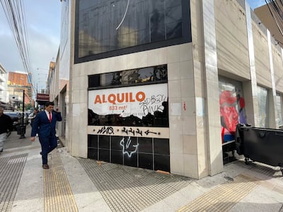 Hombre de traje azul y corbata roja camina en una calle desierta, con un cartel de 'ALQUILO' al fondo.