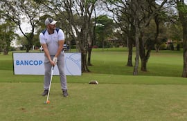 Hombre concentrado, vestido con camiseta blanca y gris, guantes y pantalones grises, en un campo de golf con un cartel de Bancop detrás.