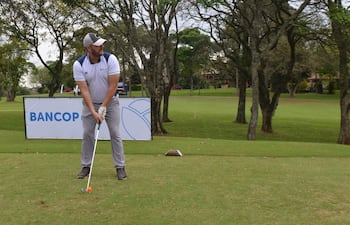 Hombre concentrado, vestido con camiseta blanca y gris, guantes y pantalones grises, en un campo de golf con un cartel de Bancop detrás.