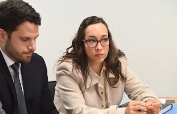 Hombre con barba y traje oscuro escucha atento, mientras mujer de cabello rizado y chaqueta clara parece reflexionar en una reunión.